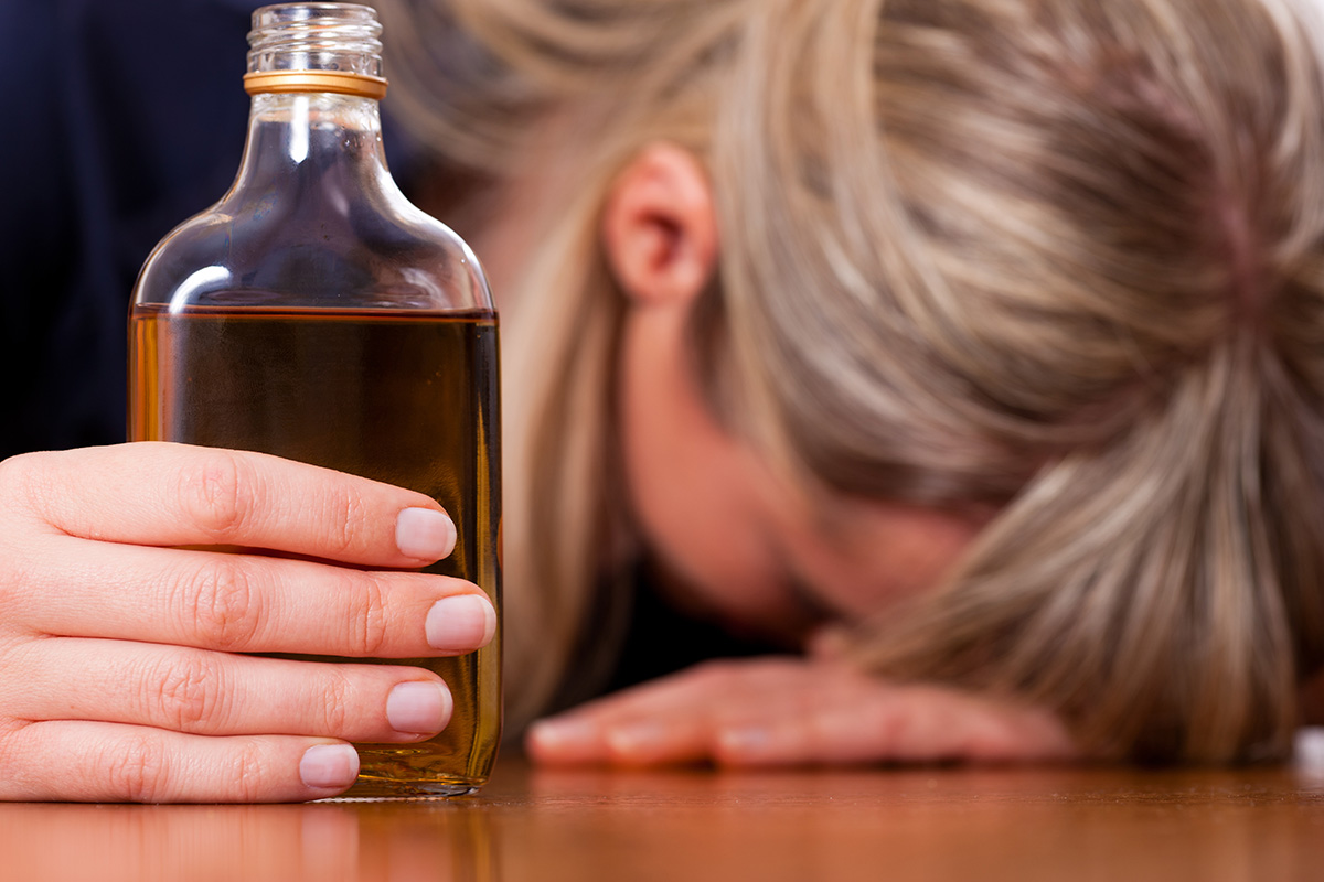 woman with head down on table holding alcohol and suffering the Biological Effects of Alcoholism
